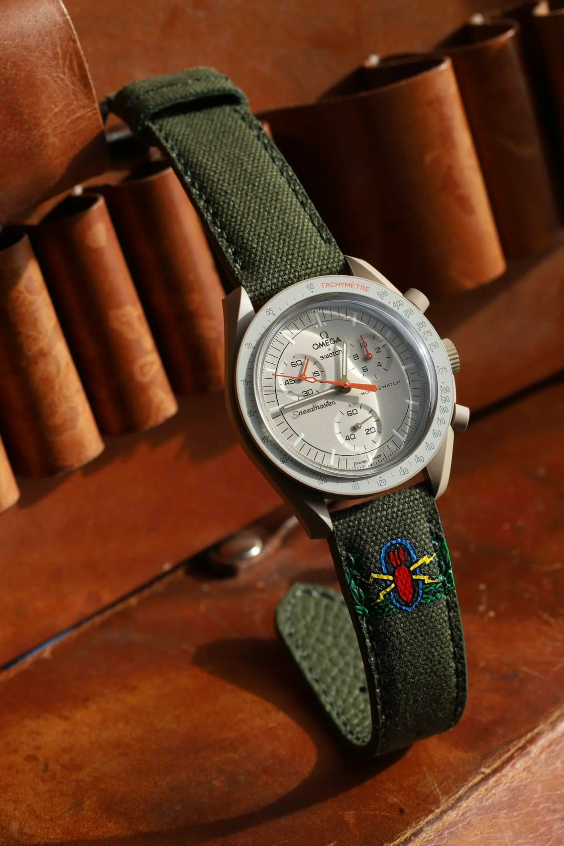 An Omega Speedmaster MoonSwatch on olive canvas with a hand-embroidered motif, resting on a leather tool roll.