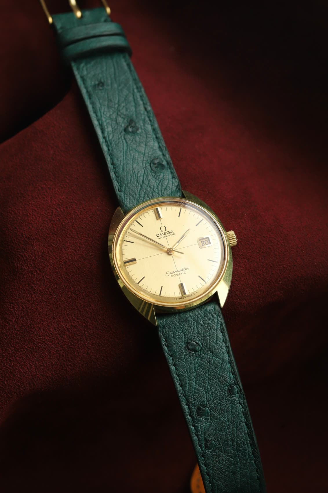 A vintage gold Omega Seamaster Cosmic on a forest-green ostrich strap, laid on burgundy suede.
